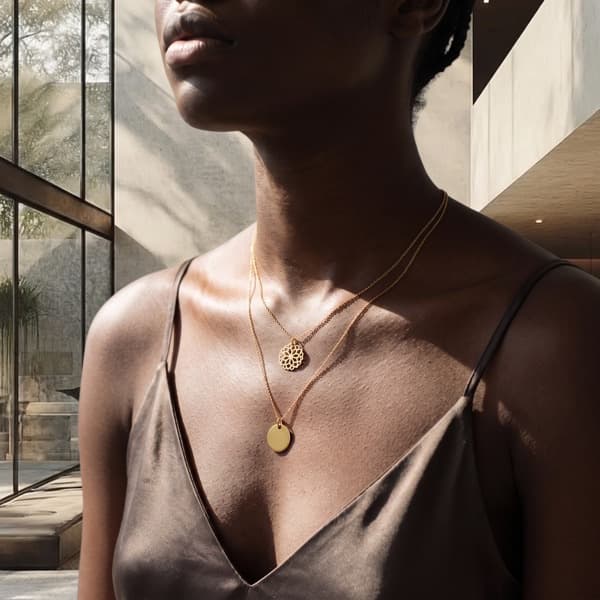 The same two necklaces worn layered at the collarbone over a slate silk camisole, sunlight falling through a glazed atrium behind.