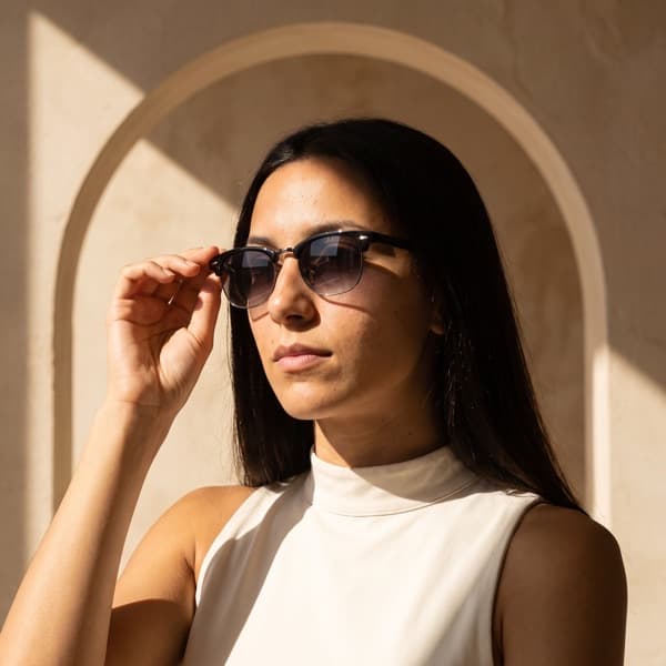 The same sunglasses worn against a cream plaster arch in hard sunlight, the model steadying one temple with her hand.