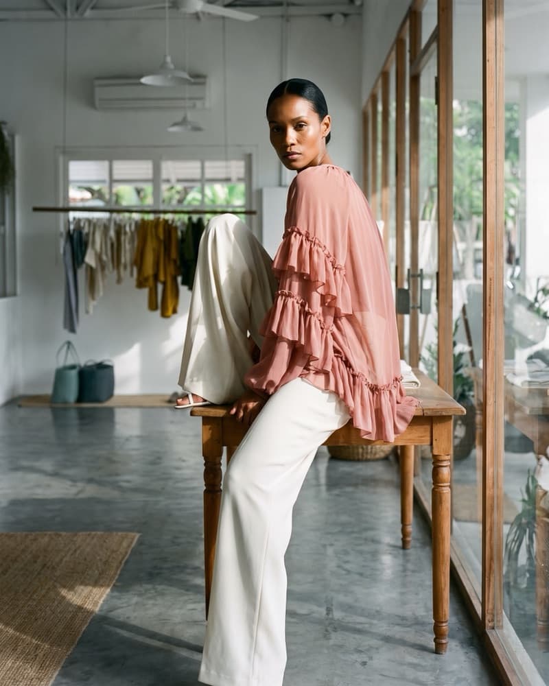 The same blouse shot full length in the boutique, the model perched on the shop's timber table and turning back to the lens.