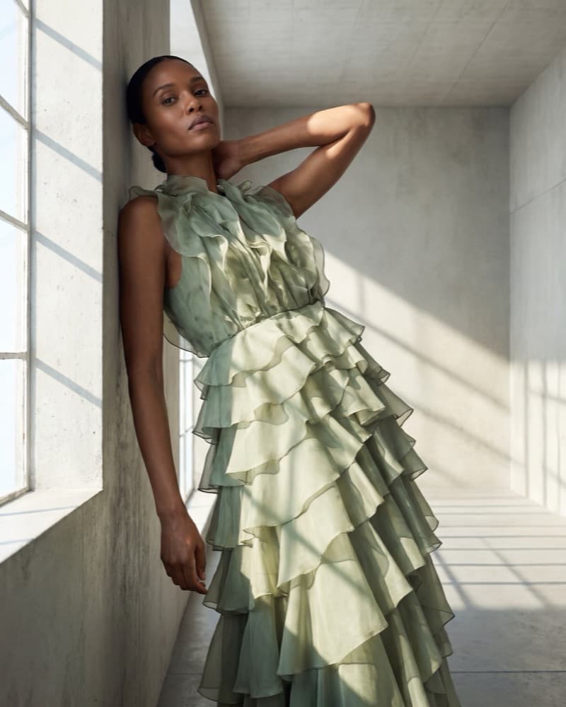 The same sage gown in a tall concrete gallery worn by one member of the cast, leaning against the window wall.